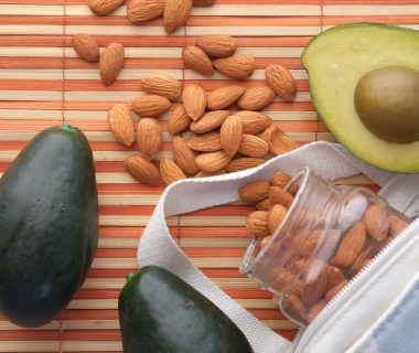 Top view of fresh avocados and almonds in a glass jar on a bamboo mat, perfect for healthy snacking.